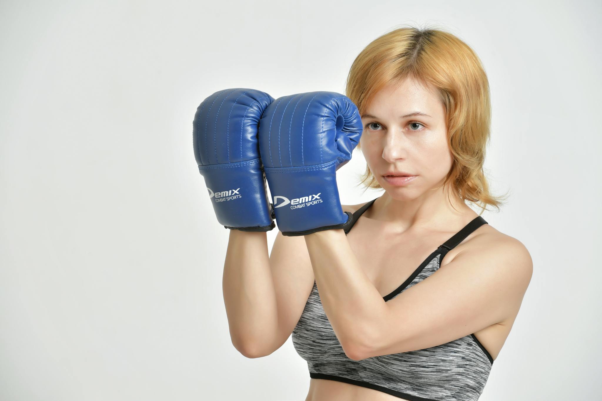 Young confident female in sports top clenching fists in blue boxing gloves on grey background